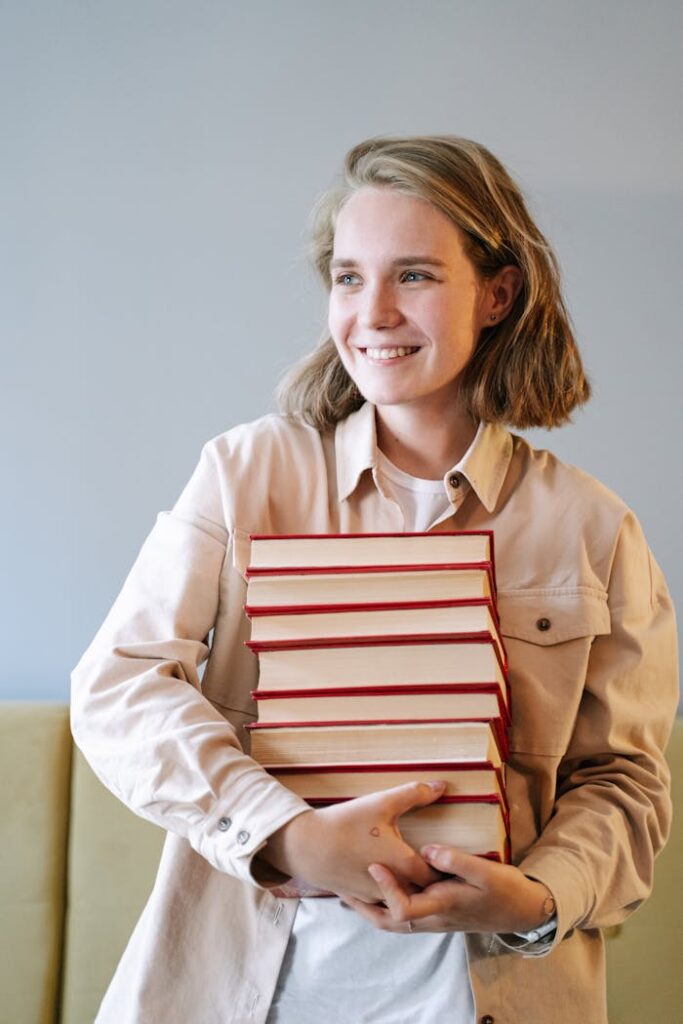 A young woman smiling while holding a stack of books indoors, embodying joy and education.